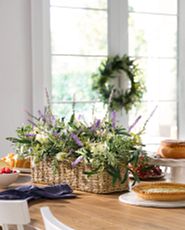 Brown dining table with an artificial flower arrangement in a rectangular basket, a bowl of mixed berries, and a cake on a white stand