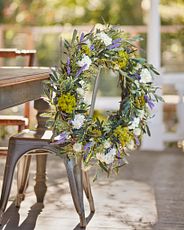 Outdoor metal dining chair decorated with artificial flower garland featuring purple cattails, white lilacs, and thistle with mixed greenery